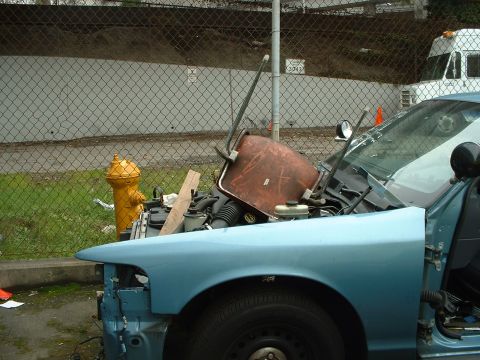 1999 Ford Crown Victoria - A light blue car with its hood open, revealing engine parts and a bucket, parked outdoors near a fence and fire hydrant.