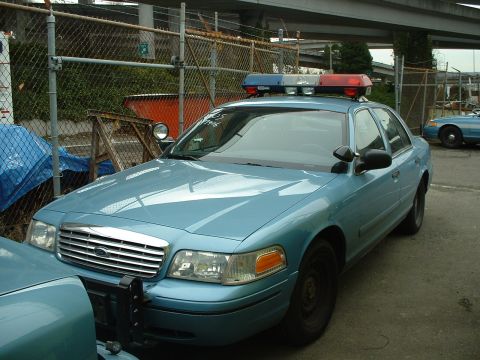 1999 Ford Crown Victoria - Two light blue police cars parked outdoors, one in the foreground with a light bar on its roof.