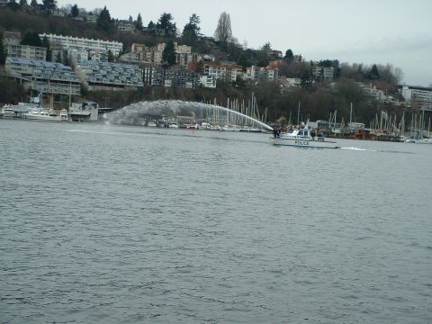 1999 Ford Crown Victoria - A police boat sprays water in a harbor, with boats docked and buildings on a hill overlooking the water.