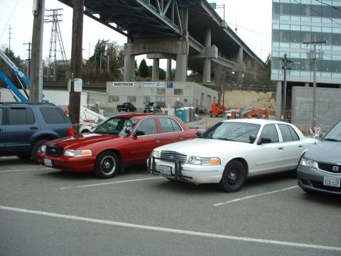 1999 Ford Crown Victoria - Two Ford Crown Victoria police cars are parked in a lot under an overpass.