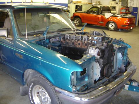 1997 Ford Ranger - A teal 1997 Ford Ranger is shown with its engine removed, revealing the engine bay, while a red Ford F-150 is parked in the background.