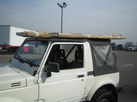 1990 Ford Bronco - white Suzuki Samurai with a soft top has a long wrapped package strapped across its roof while parked in an open lot.