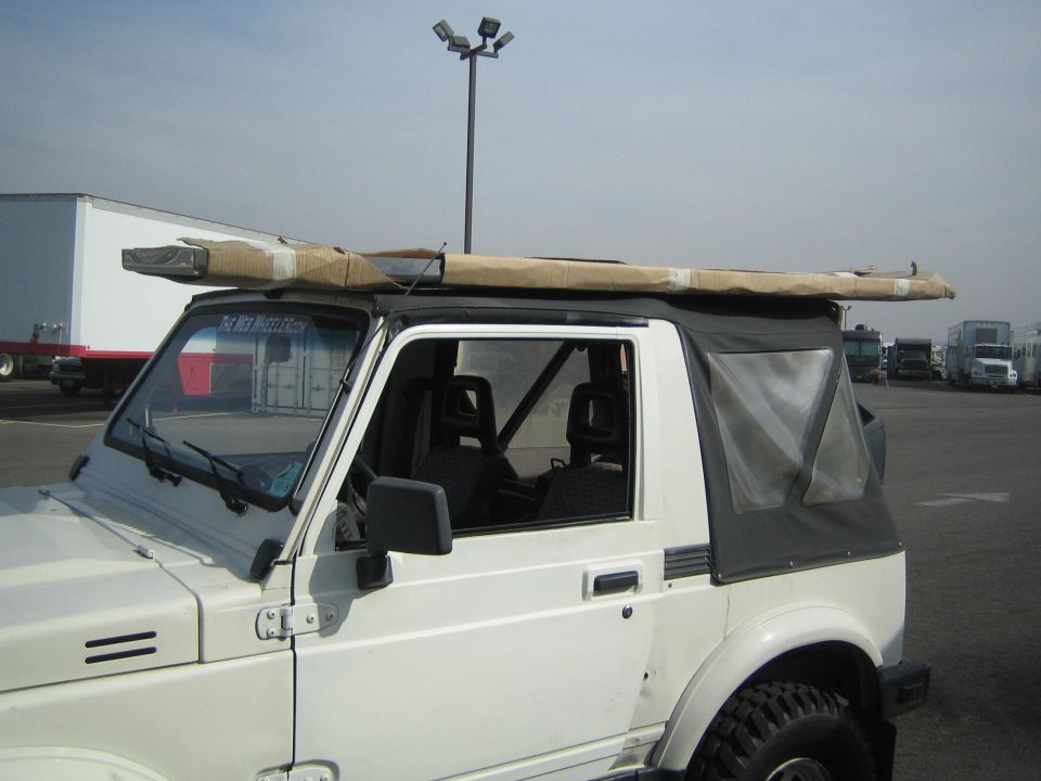 White Suzuki Samurai soft-top with long package strapped to roof in a parking lot