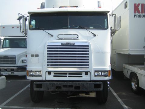 1990 Ford Bronco - white Freightliner cabover semi-tractor seen head-on, parked among box trucks and trailers in an outdoor lot.