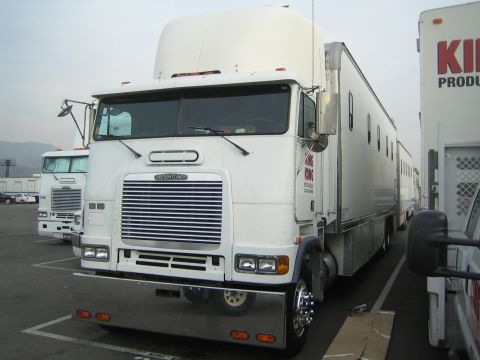 1990 Ford Bronco - white Freightliner cabover semi-truck sits parked in an asphalt lot next to box trailers and production vehicles.