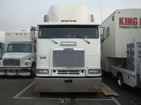 1990 Ford Bronco - Head-on view of a white Freightliner cabover semi-truck parked in a lot with trailers and a smaller truck beside it.