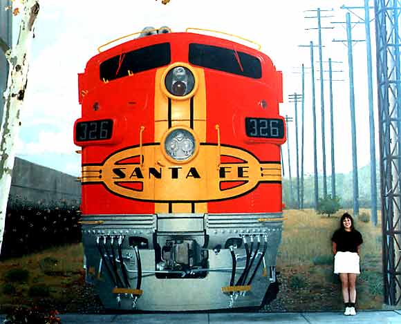 Front of a red and yellow Santa Fe train engine with '326' on the side, a woman stands to the right