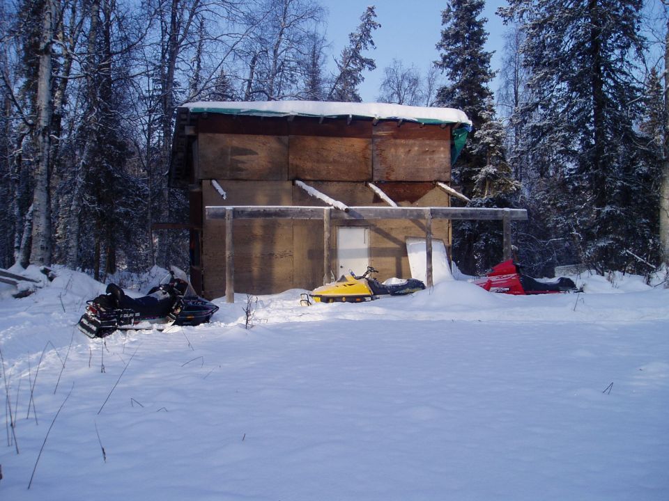 Three snowmobiles parked in the snow outside a wooden shed in a wooded area