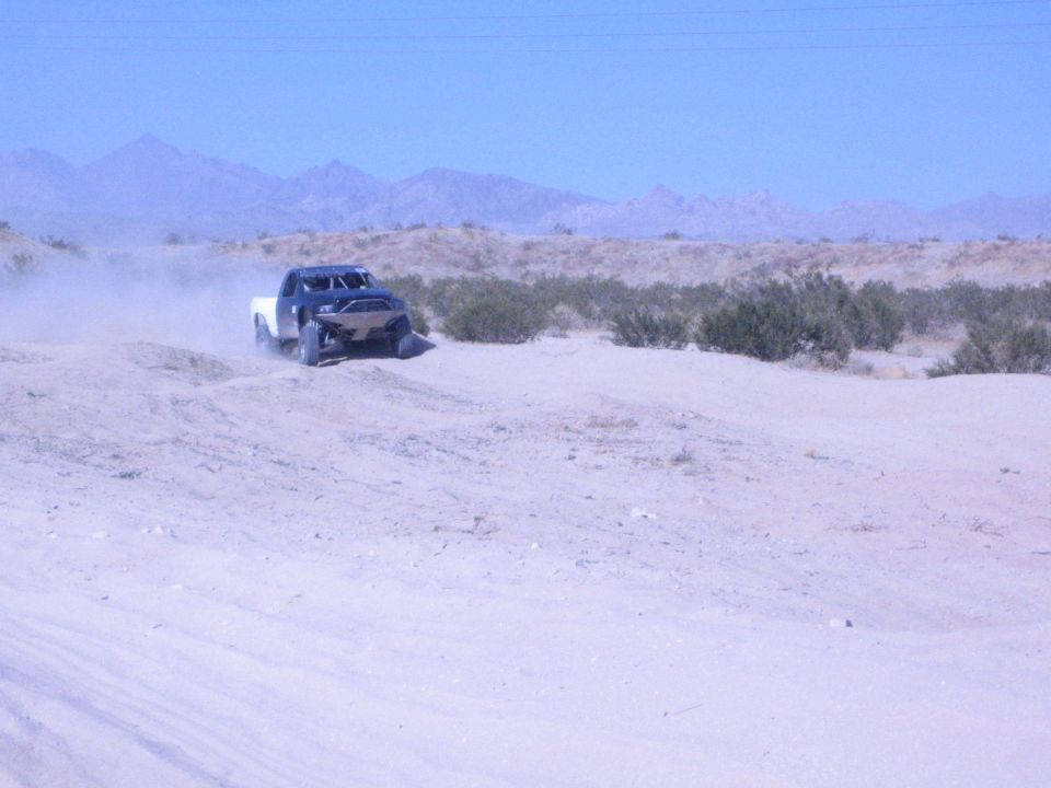 Dark off-road race truck racing across a sandy desert with mountains behind