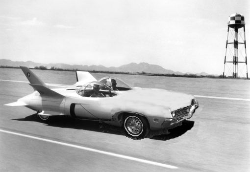 Experimental jet car with two cockpits on a paved surface, water tower in background