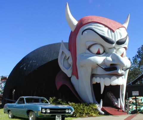 2002 Ford Crown Victoria - A vintage blue coupe is parked in front of a massive, grinning devil head roadside attraction.