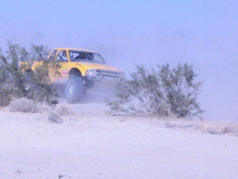 Yellow off-road race truck cutting through desert brush in heavy dust