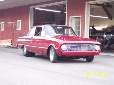 1960 Ford Falcon - A red 1960 Ford Falcon coupe hot rod is parked outside a garage with its front end facing the camera.