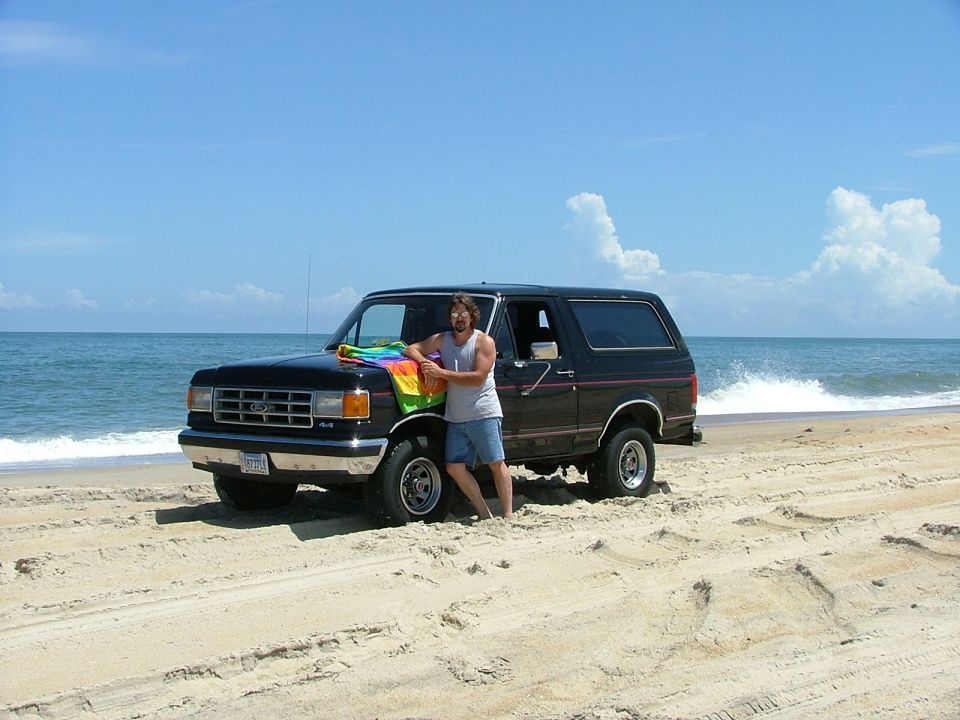 Man holding a rainbow towel leans on a black Ford Bronco at the beach