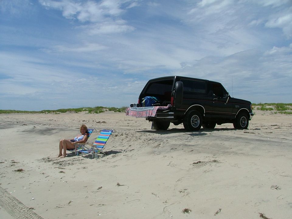 Black Ford Bronco parked on beach sand with tailgate down; woman relaxing in beach chairs nearby