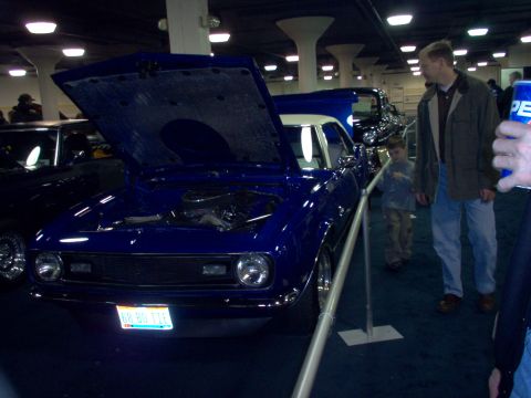 1970 Ford Ranchero - A blue 1967 Chevrolet Camaro is displayed with its hood open at an indoor event, with other vehicles visible in the background.