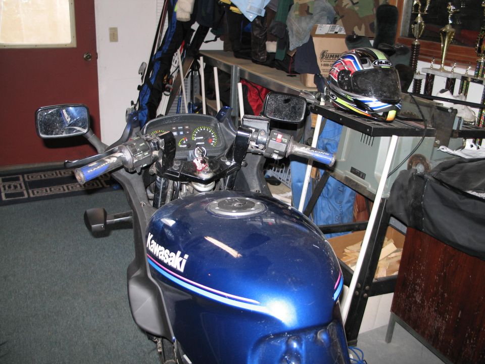 Cockpit view of blue Kawasaki motorcycle indoors, gauges and mirrors, helmet and trophies behind
