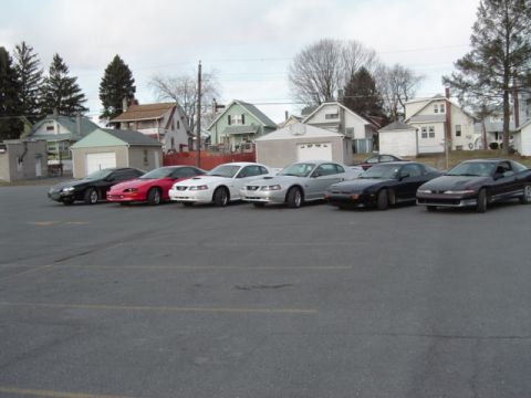 2002 Chevrolet Camaro - A line of sports cars, including red and white Ford Mustangs, are parked in a lot with houses in the background.