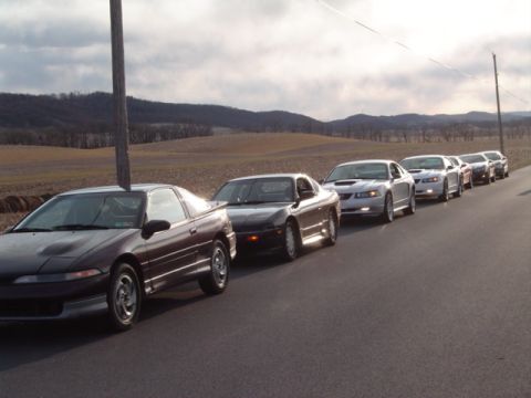 2002 Chevrolet Camaro - A line of coupes drives down a rural road under an overcast sky.