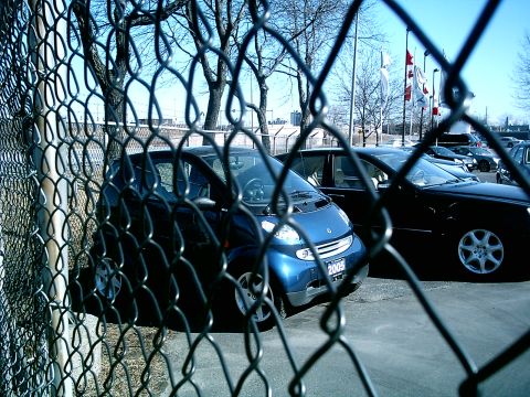 1997 Ford Escort - A blue smart car is parked behind a chain-link fence, with other vehicles visible in the background.