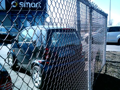 1997 Ford Escort - A dark blue Smart car is parked behind a chain-link fence, with a white SUV in the background.