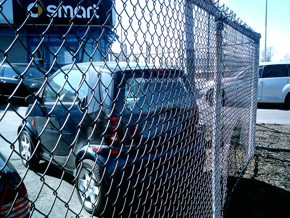 Dark blue Smart car, rear view, behind chain-link fence. Another white SUV visible