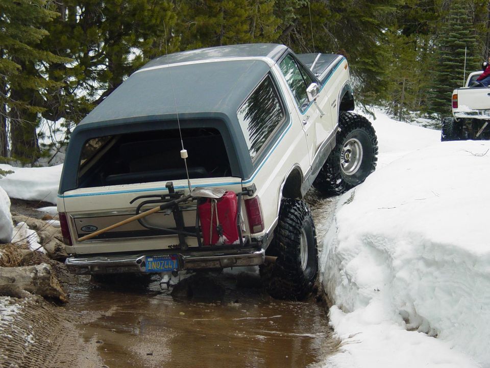 White Ford Bronco with blue trim, off-roading in snow and mud. It has a bike rack with gas cans and a shovel attached to the rear