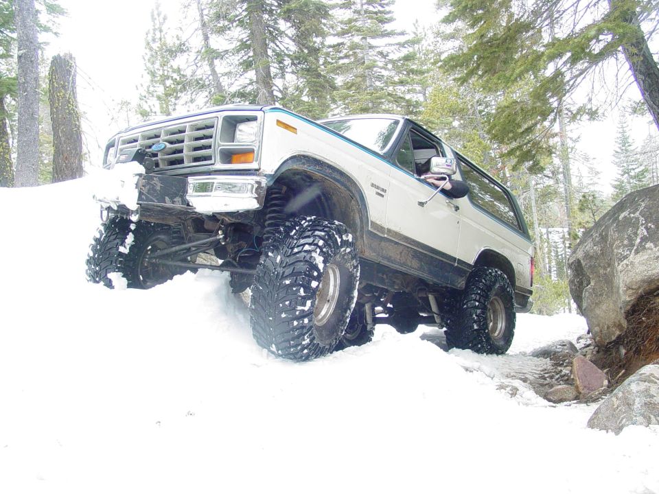1986 Ford Bronco with large tires driving through snow near rocks and trees