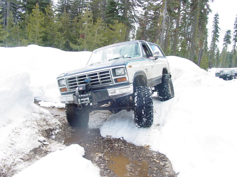Ford Bronco driving through a snowy, muddy trail with trees in the background. Another vehicle is visible further down the trail
