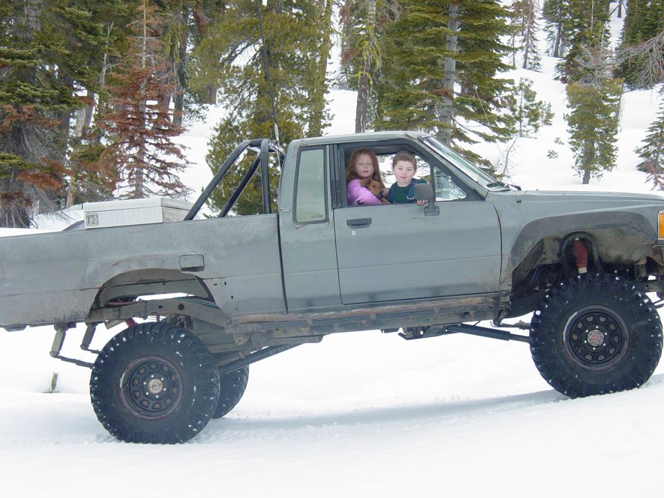 Two kids sit in a lifted, gray pickup truck with large tires in a snowy wooded area