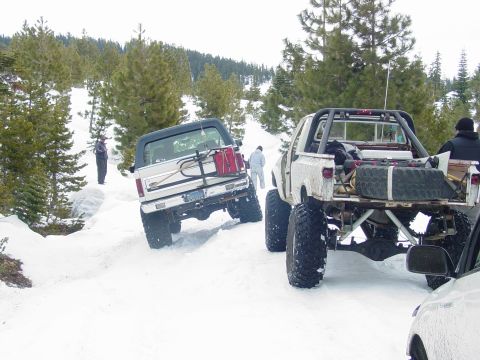 1986 Ford Bronco - Two off-road vehicles navigate a snowy trail with pine trees in the background.