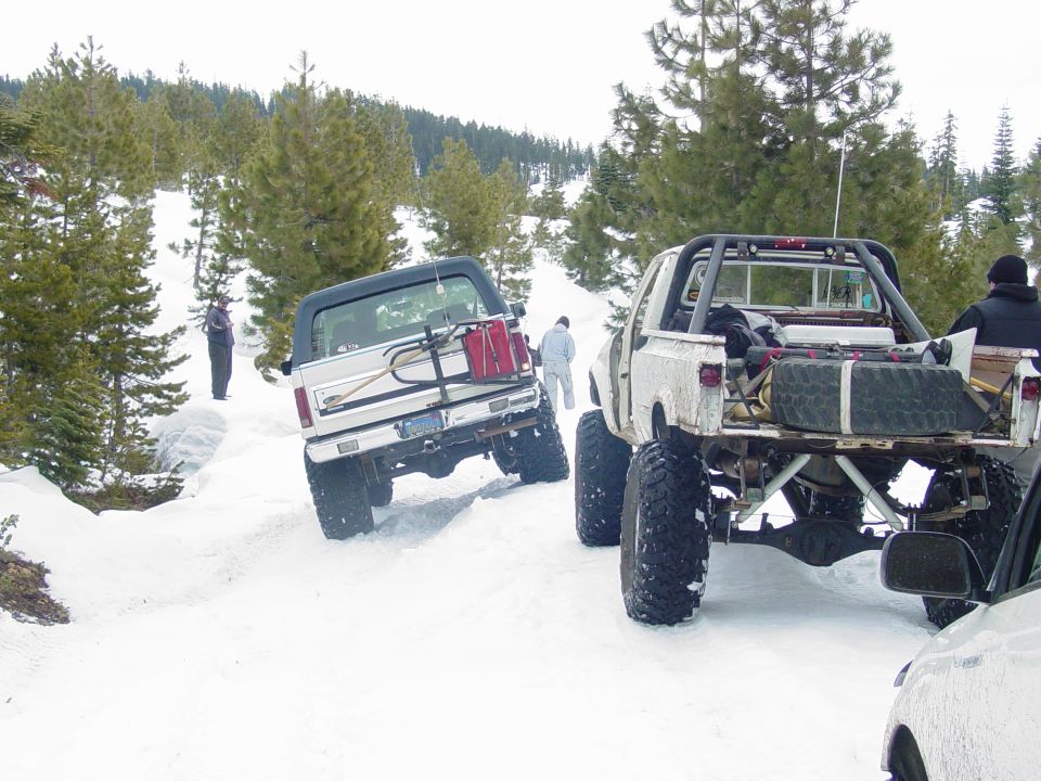 Silver Ford Bronco with a spare tire carrier and red gas cans on the back, driving in snow