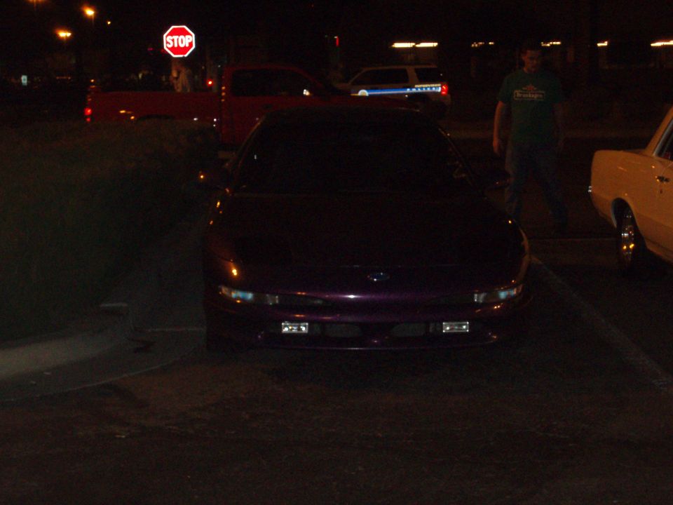 Dark purple coupe parked at night near a stop sign with a pickup and pedestrian in background