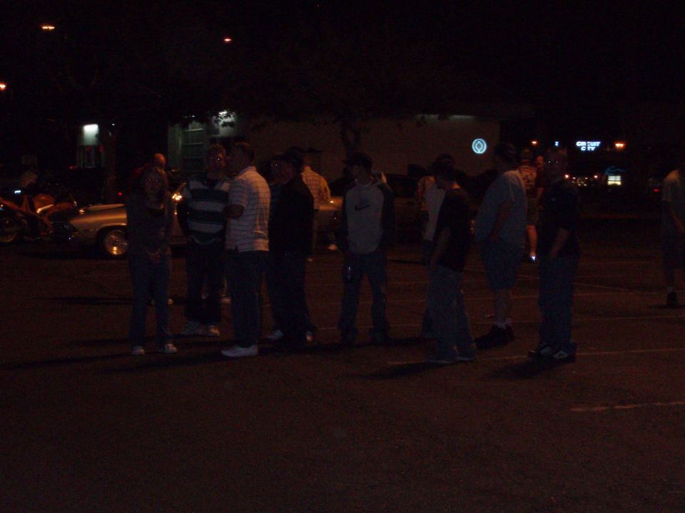 Group of people gathered around cars in a parking lot at night. A motorcycle is visible on the left