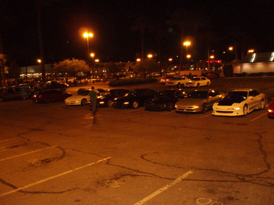 Several cars parked in a lot at night, including a white car with a black hood