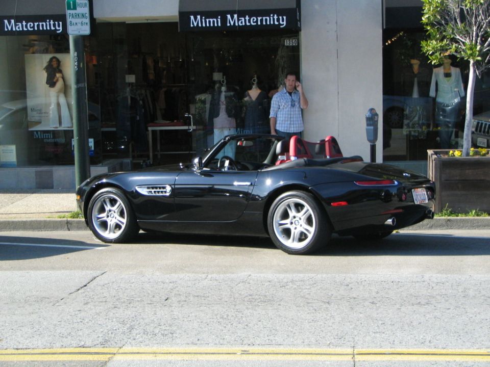 Black convertible parked on street in front of Mimi Maternity store. Man in plaid shirt talks on phone