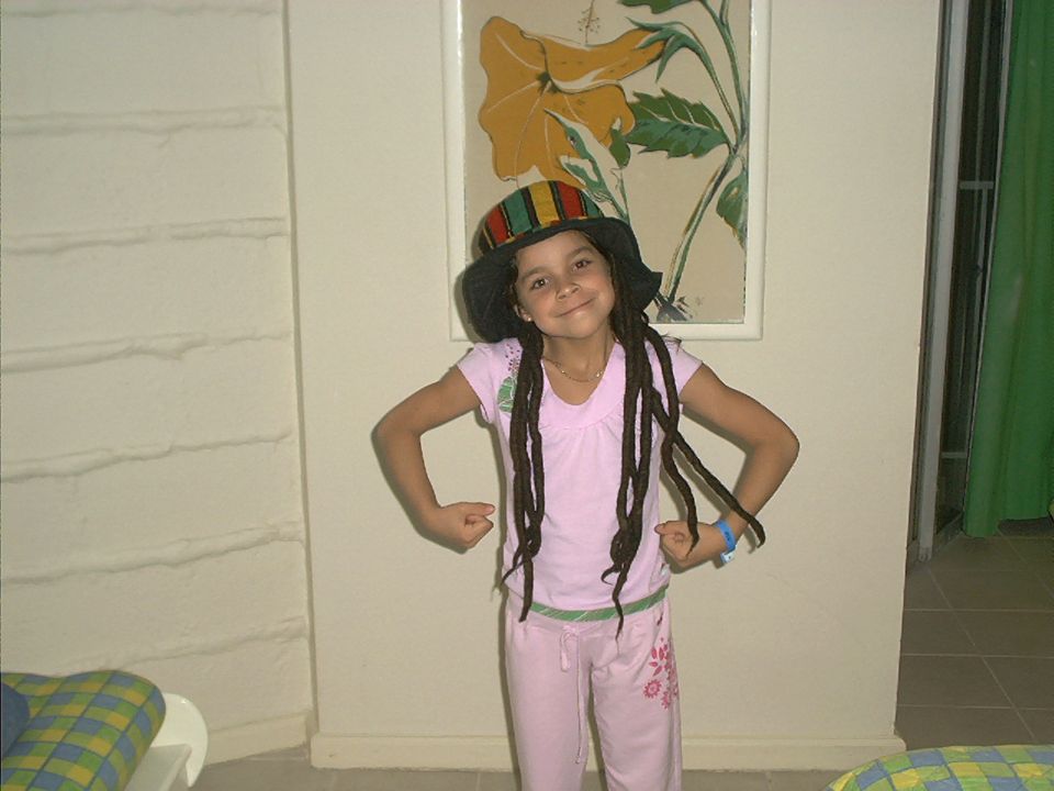 Child in pink outfit and rasta-style novelty hat posing indoors