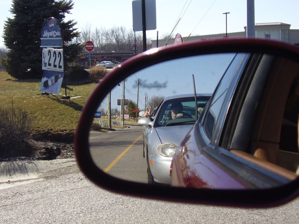 Side mirror view of a silver sedan in traffic. Driver is visible. A Pepsi sign is in the background