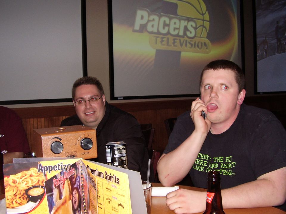 Two men sit at a table in front of a projection screen displaying 'Pacers Television'. One man talks on a cell phone with a surprised expression
