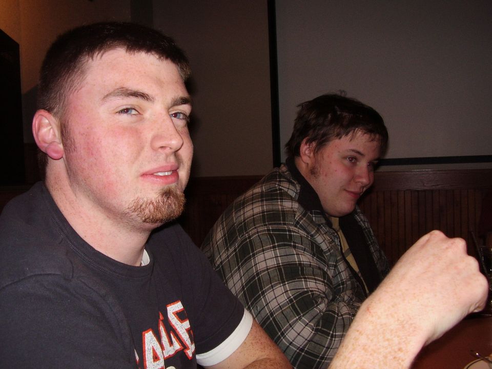 two guys sitting at a table, one in foreground wearing black shirt, other in background wearing plaid shirt