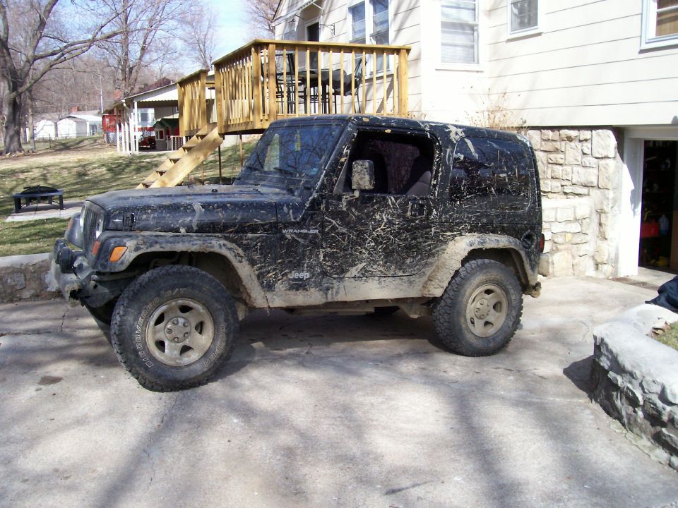 Mud-caked black Jeep Wrangler on oversized tires parked in a driveway by a wood deck