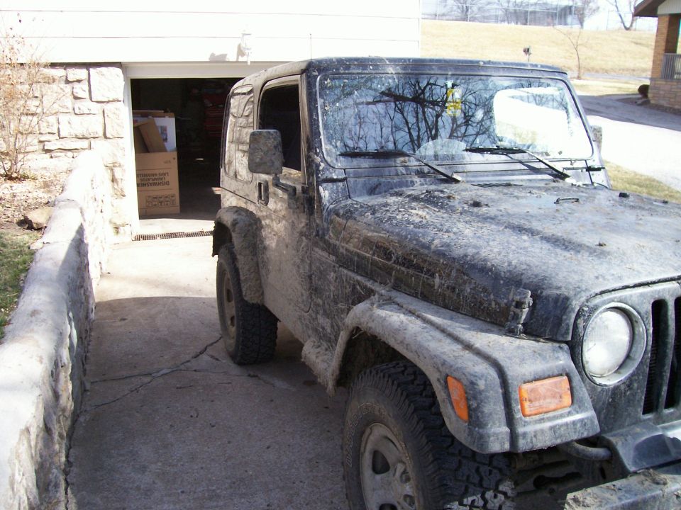 Mud-covered Jeep Wrangler parked in a driveway next to a garage