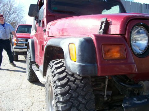 1998 Jeep TJ - A red 1998 Jeep TJ is shown with its front fender and large off-road tire prominently displayed, with another vehicle in the background.