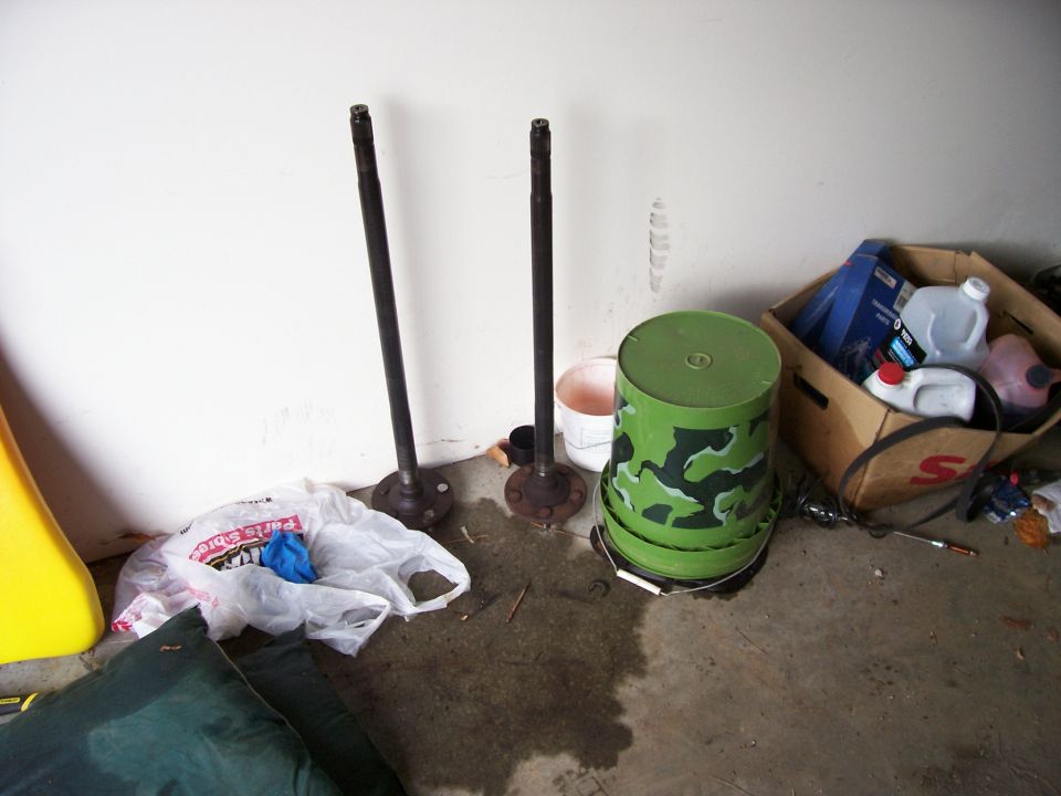 Two long axle shafts standing upright against a garage wall beside a camo bucket and supply box