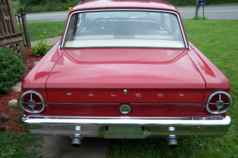 1965 Ford Falcon - The rear of a red 1964 Ford Falcon is shown, parked on a grassy lawn with a wooden fence in the background.