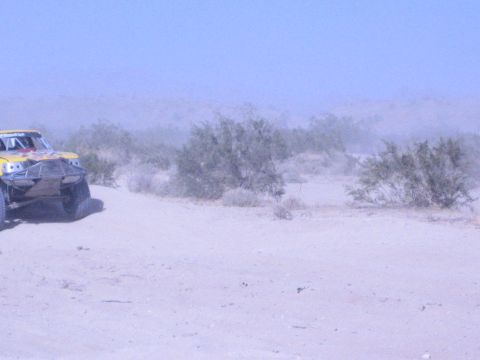 Yellow off-road race truck at left amid desert brush and dust
