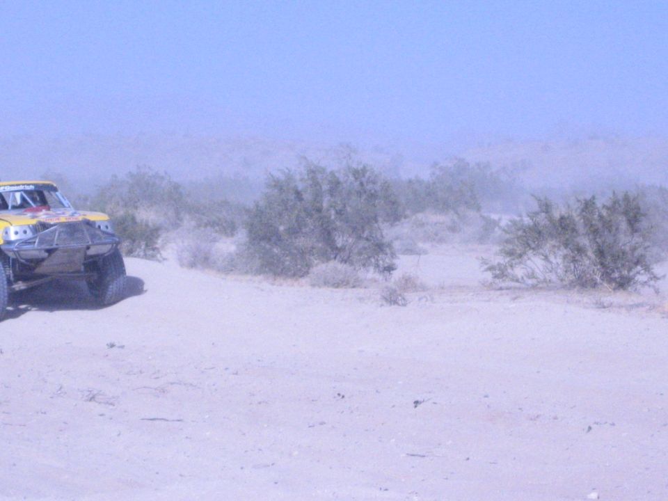 Yellow off-road race truck at left amid desert brush and dust