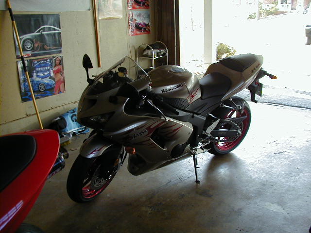 Silver Kawasaki Ninja motorcycle parked in garage next to a red motorcycle. "Kawasaki" and "Ninja" text visible