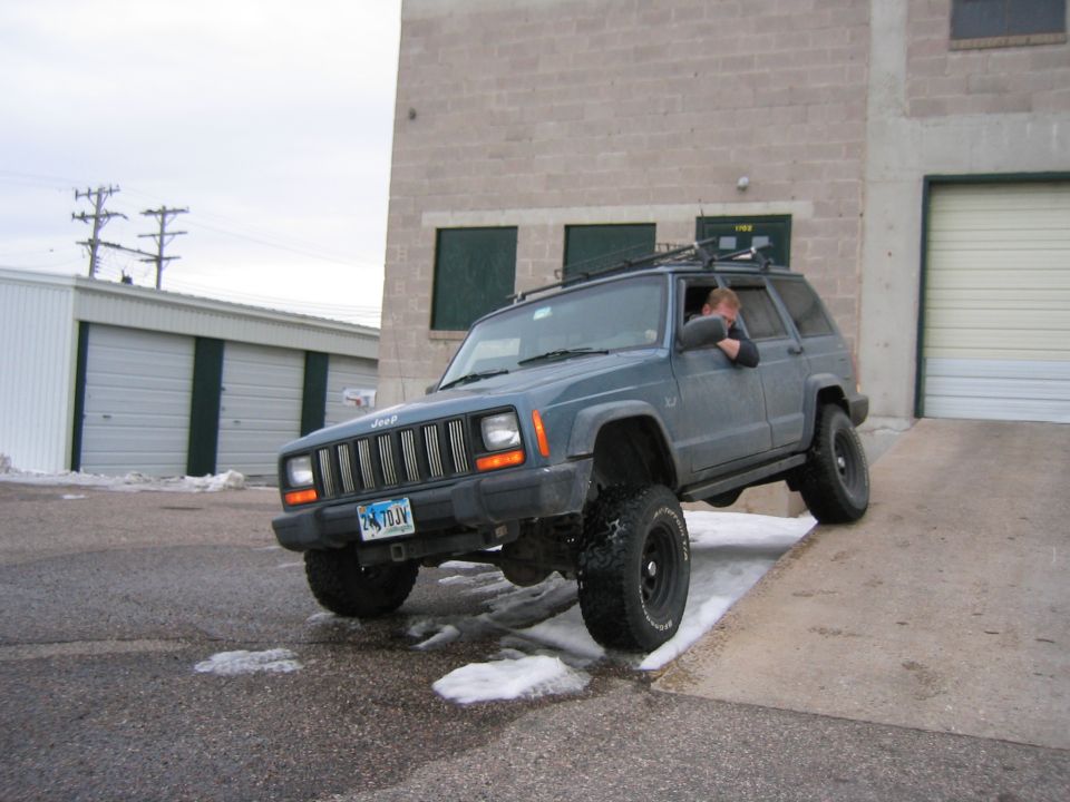 Blue-gray Jeep Cherokee XJ flexing off a concrete ramp outside a shop, driver leaning out window