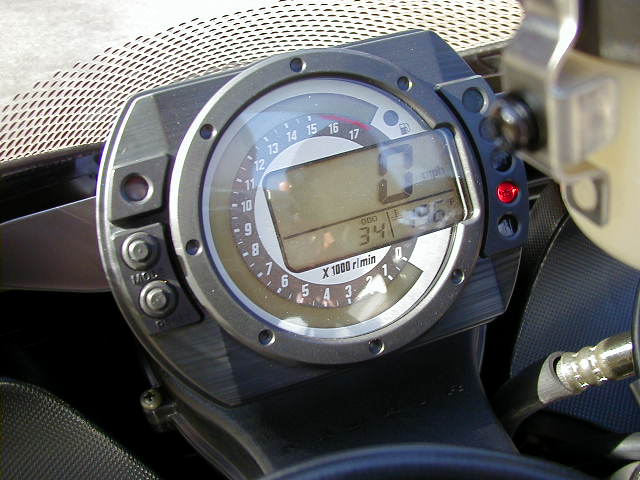 Motorcycle dashboard with digital speedometer showing 0 mph and tachometer reading 34 x 1000 r/min. Red indicator light is visible